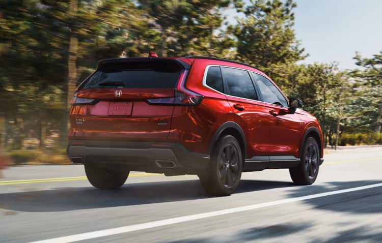 Passenger rear view of 2025 CR-V Sport AWD in Radiant Red Metallic driving down tree-lined road, kicking up dust and leaves