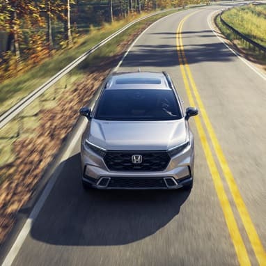 2027 Honda CR-V Sport Touring Hybrid in Solar Silver Metallic top driving on a winding road through a forest (Compact Crossover SUV).
